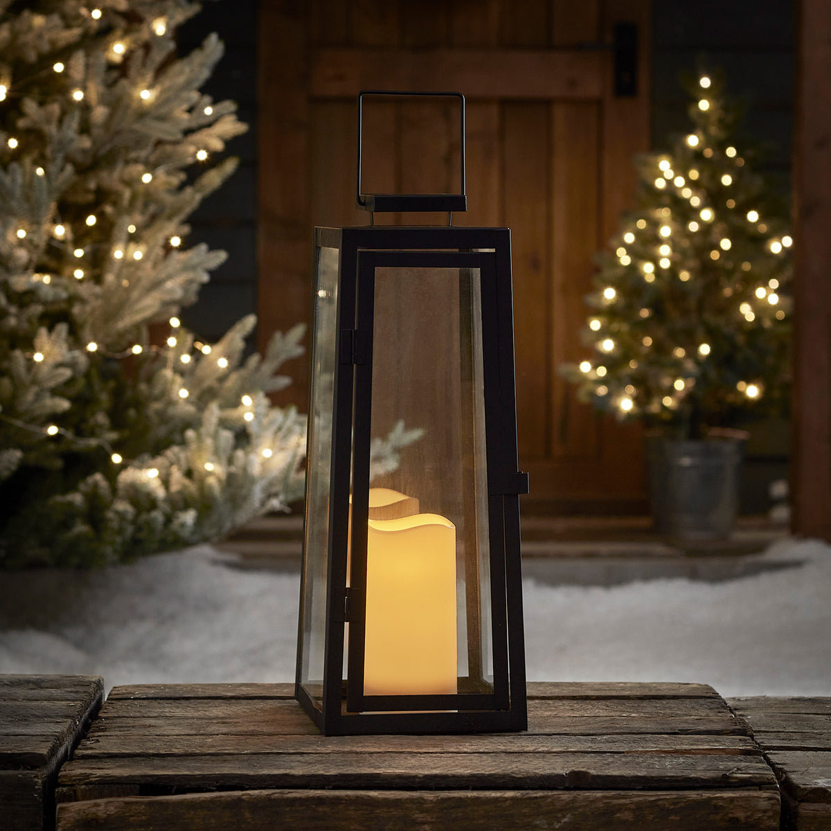 Solar-Gartenlaterne mit LED-Kerze auf Holzbrett vor beleuchteten Weihnachtsbäumen