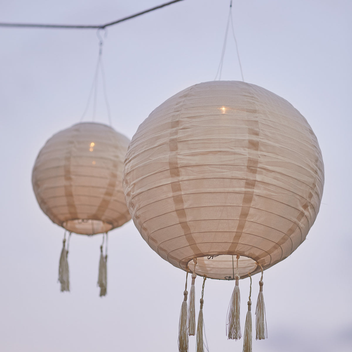 Zwei cremefarbene Solar-Lampion mit Fransen hängen bei Dämmerung an einer Schnur
