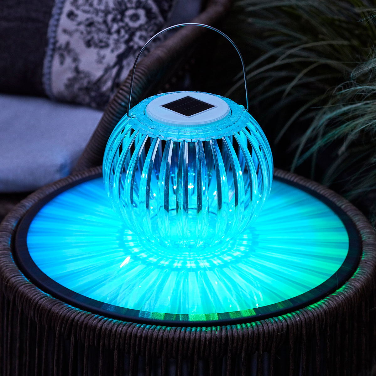 Solar Hängeleuchte leuchtet in blauem Farbton auf Gartentisch mit Pflanzen im Hintergrund
