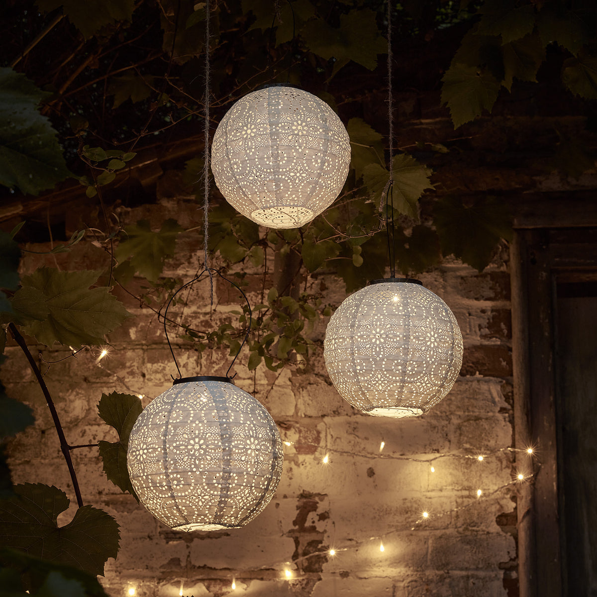 Drei weiße, beleuchtete Tyvek Solar Lampions mit Blumenmuster hängen im Garten vor einer Backsteinwand