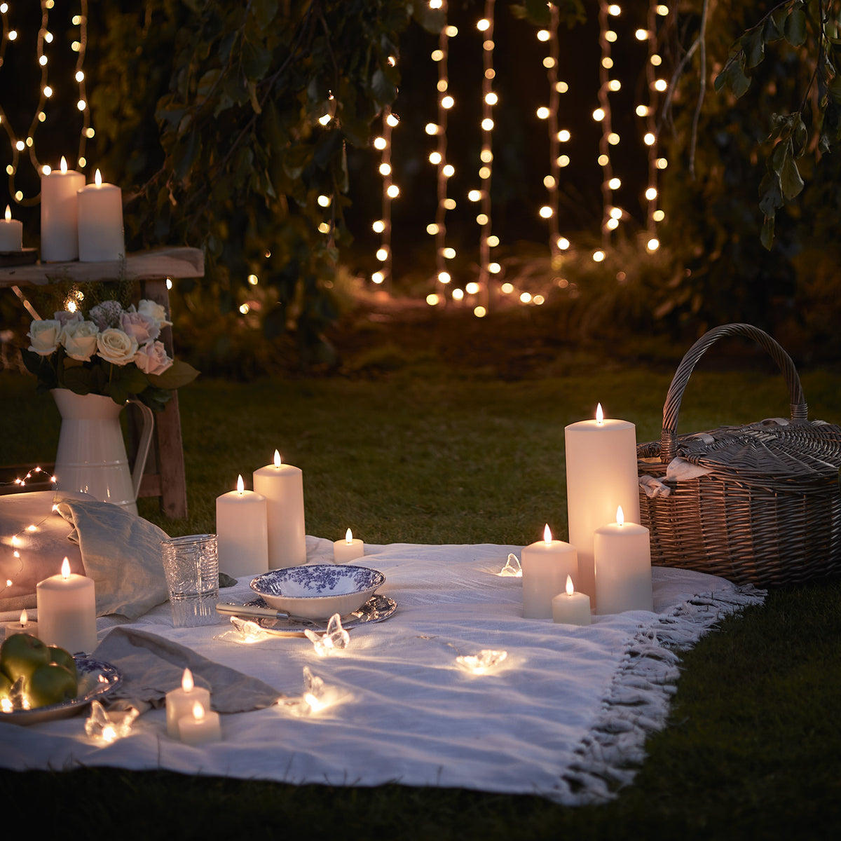 Picknickdecke mit mehreren brennenden TruGlow LED Kerzen und Lichterketten im Garten