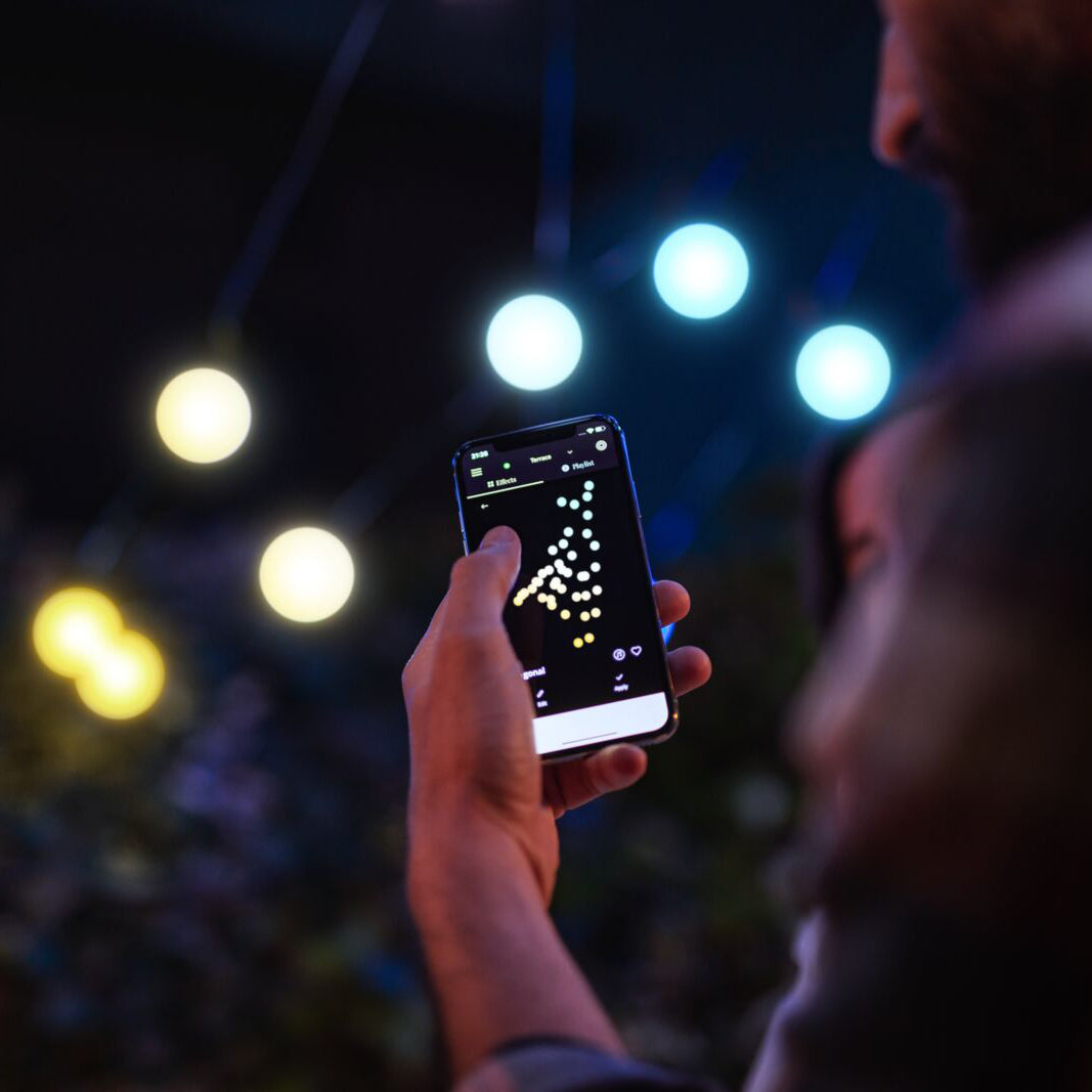 Person steuert smarte Lichterkette mit Smartphone-App, LED-Glühbirnen leuchten orange und weiß