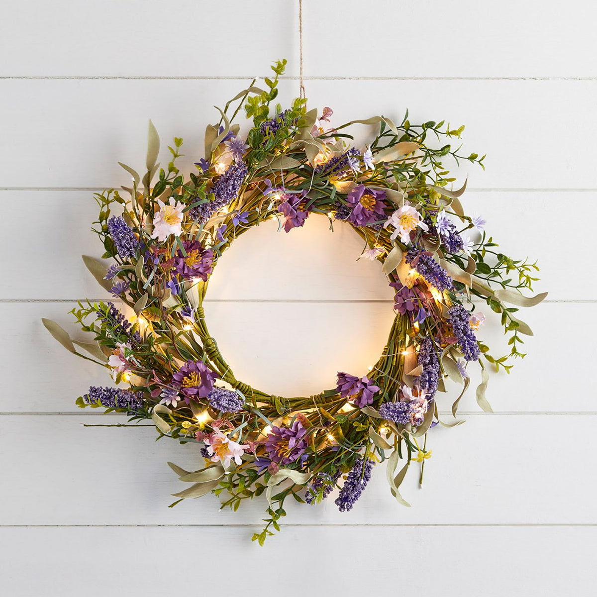 Lavendelkranz mit bunten Blumen und Licht an Kabel vor weißer Wand.