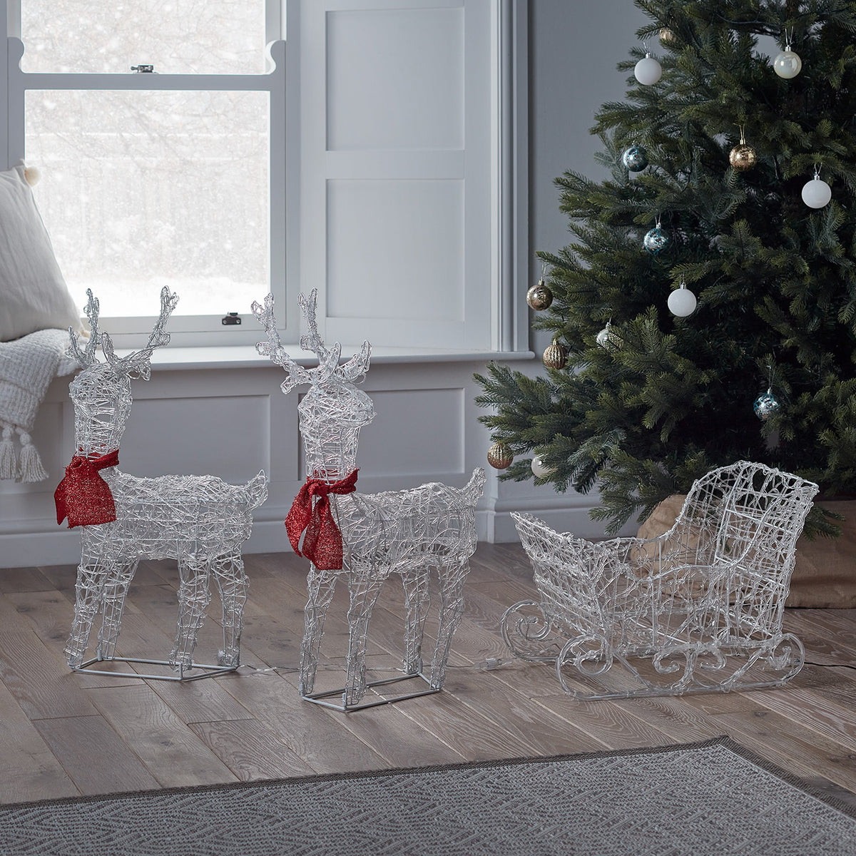 Unbeleuchtete Acryl-Rentiere mit roten Schals und ein Schlitten neben geschmücktem Weihnachtsbaum