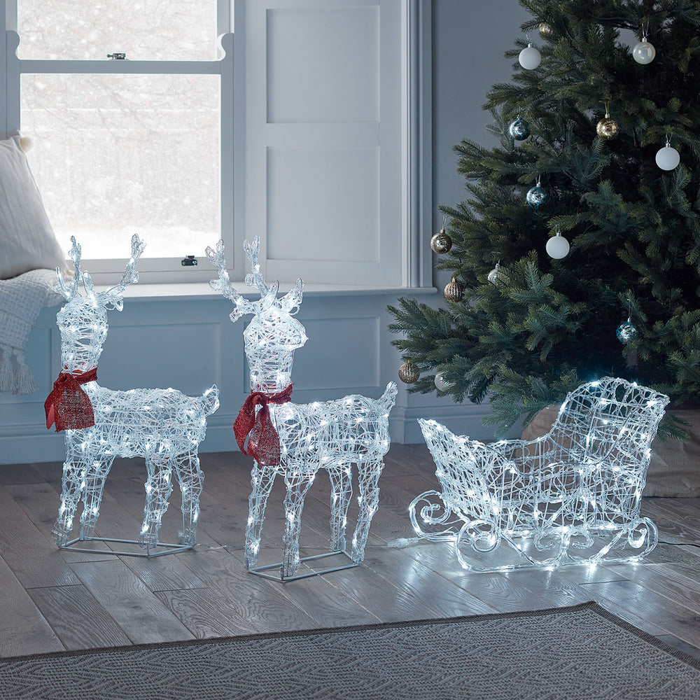 Zwei batteriebetriebene LED-Rentiere mit roten Schals und ein beleuchteter Schlitten vor Fenster und Weihnachtsbaum