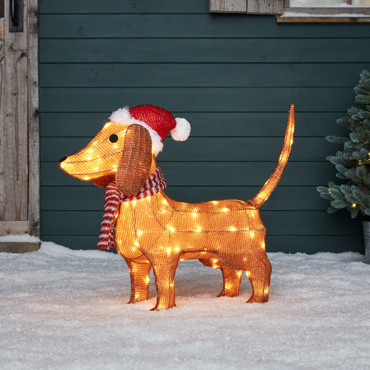 Leuchtende LED Dackel Weihnachtsfigur mit roter Mütze und Schal steht im Schnee vor grünem Holzwand