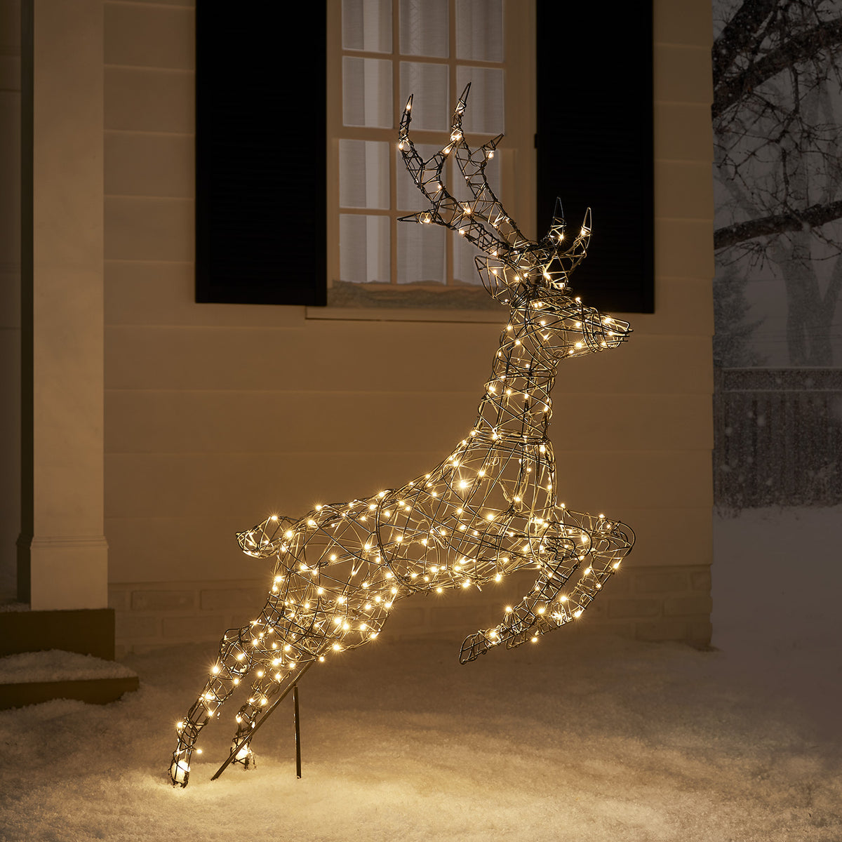 Springendes LED-Rentier mit warmweißen Lichtern vor Hausfassade im Schnee