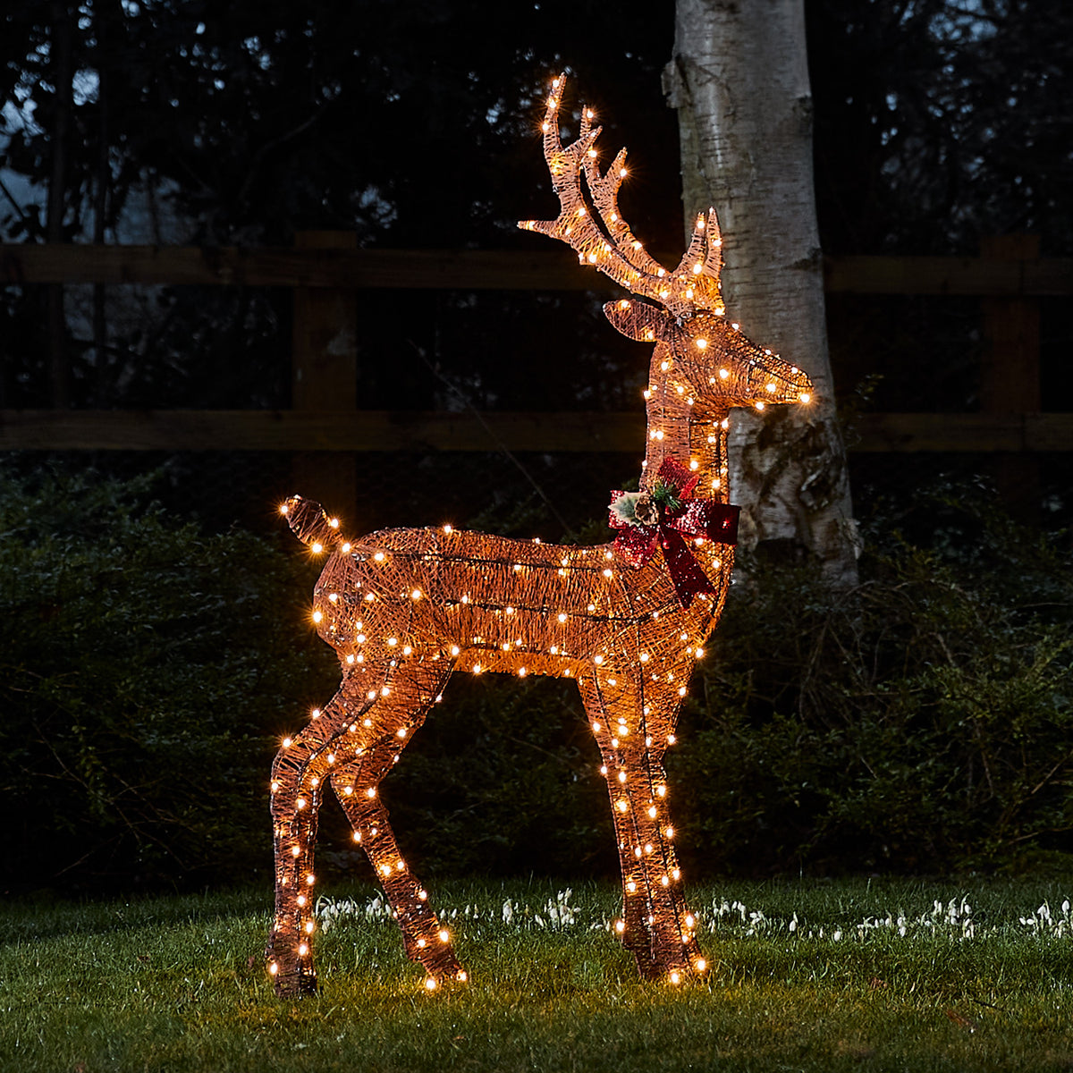 Braunes LED-Rentier mit roter Schleife leuchtet nachts im Garten vor Baum